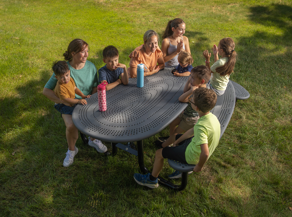 The Community Table site furnishings with integrated infant seat, showing a multi-generational gathering in a park setting.